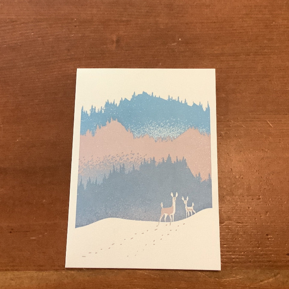 A small art print or postcard featuring a stylized winter landscape with blue and pink mountains, dark trees, and two deer in the snow, placed on a wooden tabletop.