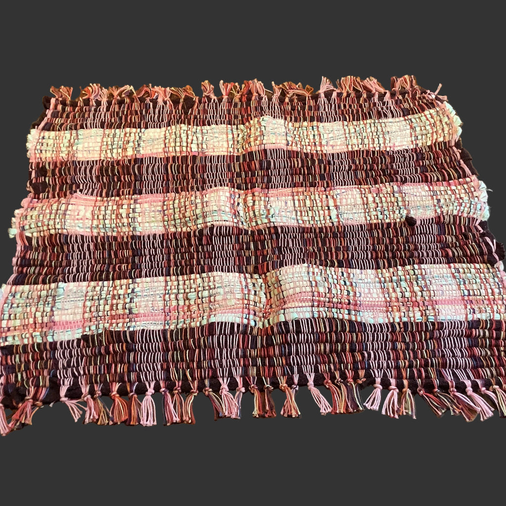 A rectangular woven placemat with fringed edges, featuring horizontal stripes in burgundy, pink, and cream, laid flat on a dark wood table.