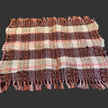 A rectangular woven placemat with fringed edges, featuring horizontal stripes in burgundy, pink, and cream, laid flat on a dark wood table.