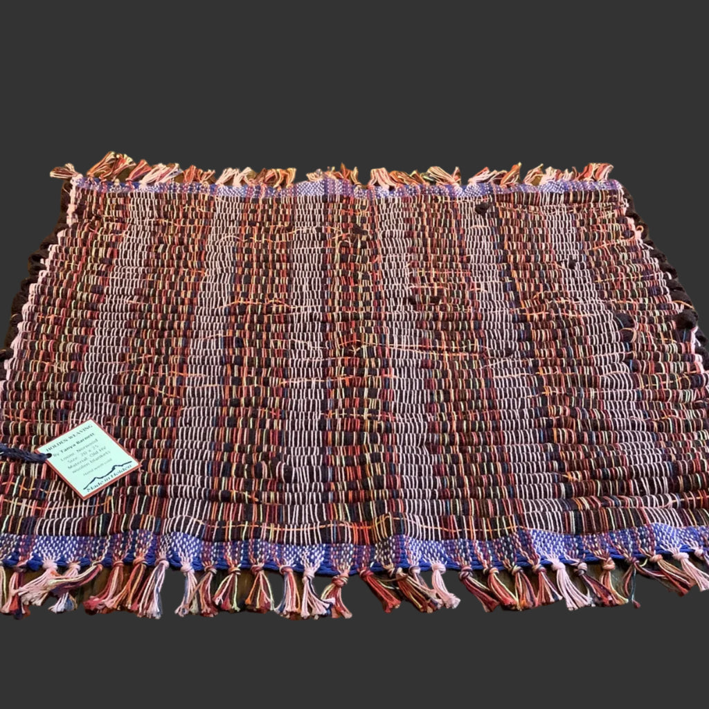 A rectangular woven textile placemat with fringe edges, laid flat on a dark wood table in front of a light wood slat wall. The placemat has a multicolor pattern with vertical stripes in burgundy, red, purple, pink, and cream, with a blue-purple patterned border.
