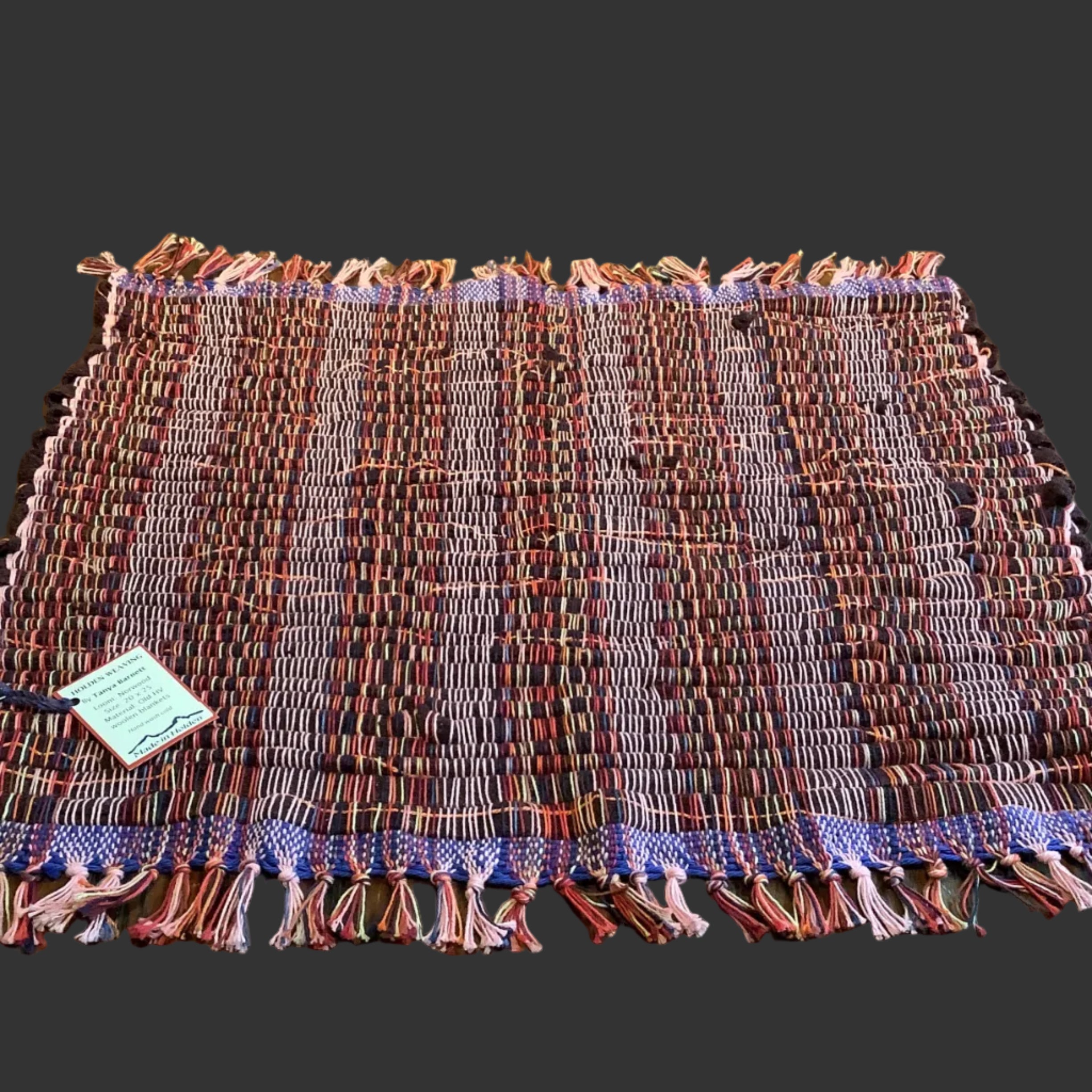 A rectangular woven textile placemat with fringe edges, laid flat on a dark wood table in front of a light wood slat wall. The placemat has a multicolor pattern with vertical stripes in burgundy, red, purple, pink, and cream, with a blue-purple patterned border.