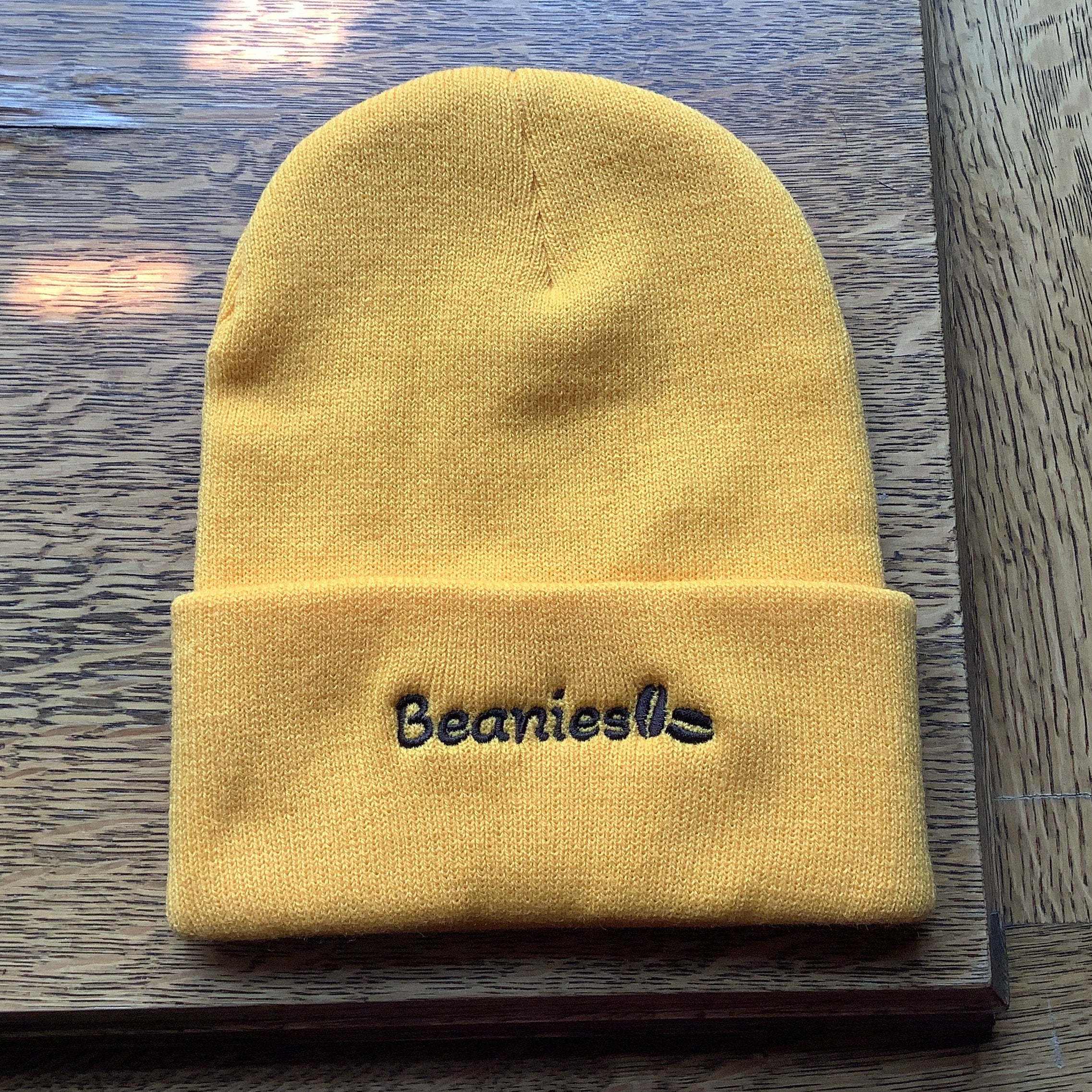 A yellow knit beanie with a folded cuff featuring the embroidered text 'Beanies' in black, resting on a dark wooden surface.