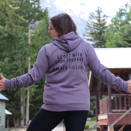 Person wearing a purple hoodie with text, standing outdoors with trees and a cabin in the background