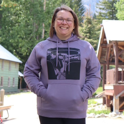 Person wearing a purple hoodie with a graphic design, standing outdoors with trees and a building in the background.