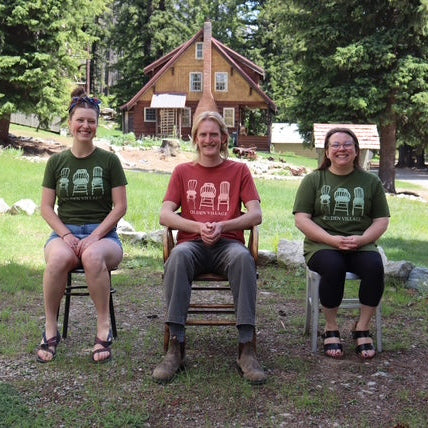 Three people sitting on chairs in front of a cabin with trees in the background