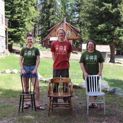 Three people standing outdoors with chairs in front of a cabin.