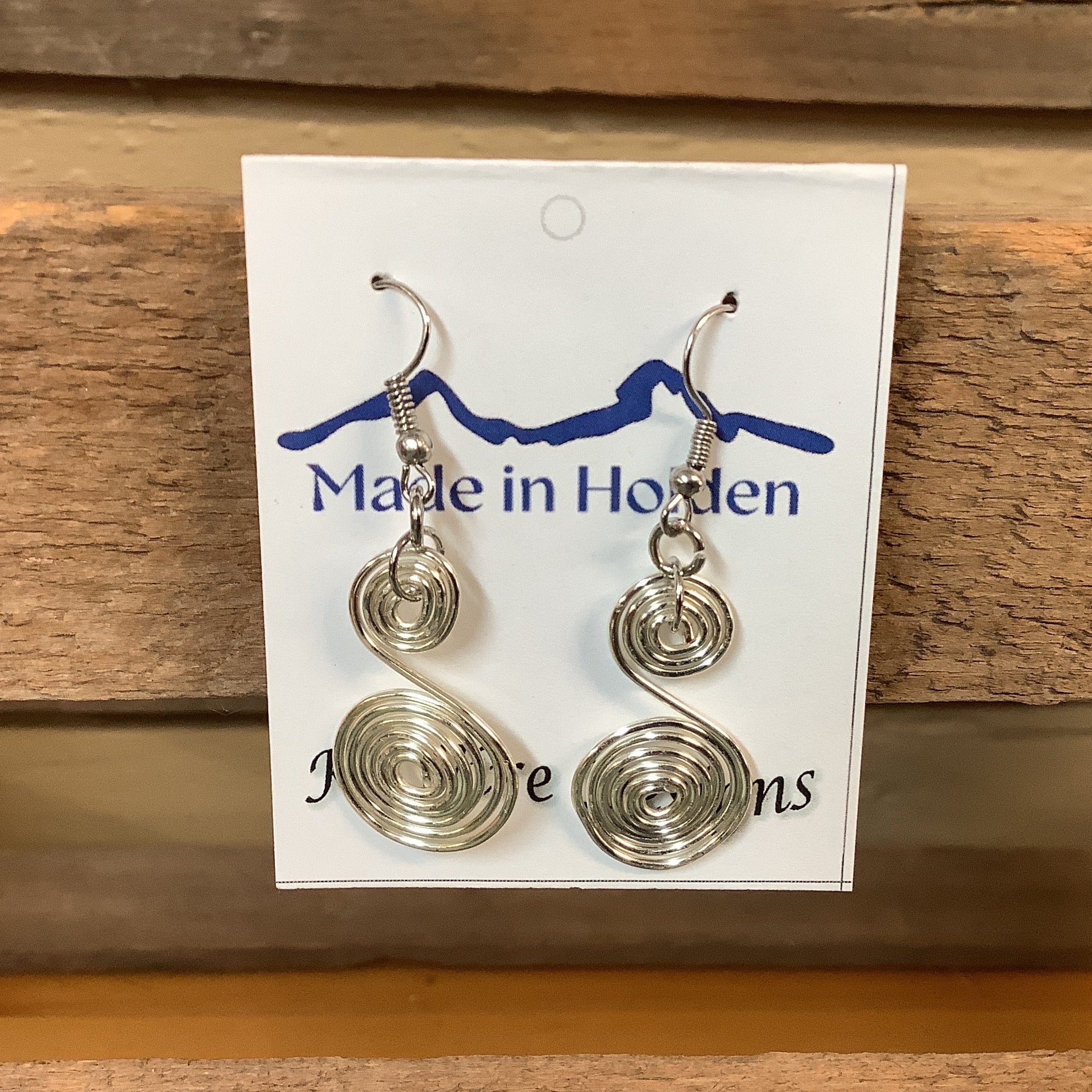 A pair of handmade silver wire spiral dangle earrings displayed on a white backing card that reads 'Made in Holden' and 'K3 Wire Designs,' hanging on a rustic wooden shelf in an indoor setting. SKU 8084