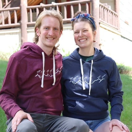 Two people wearing matching hoodies sitting on grass in front of a house.