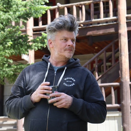 Man wearing a black hoodie with a logo, standing outdoors near wooden structures and trees.
