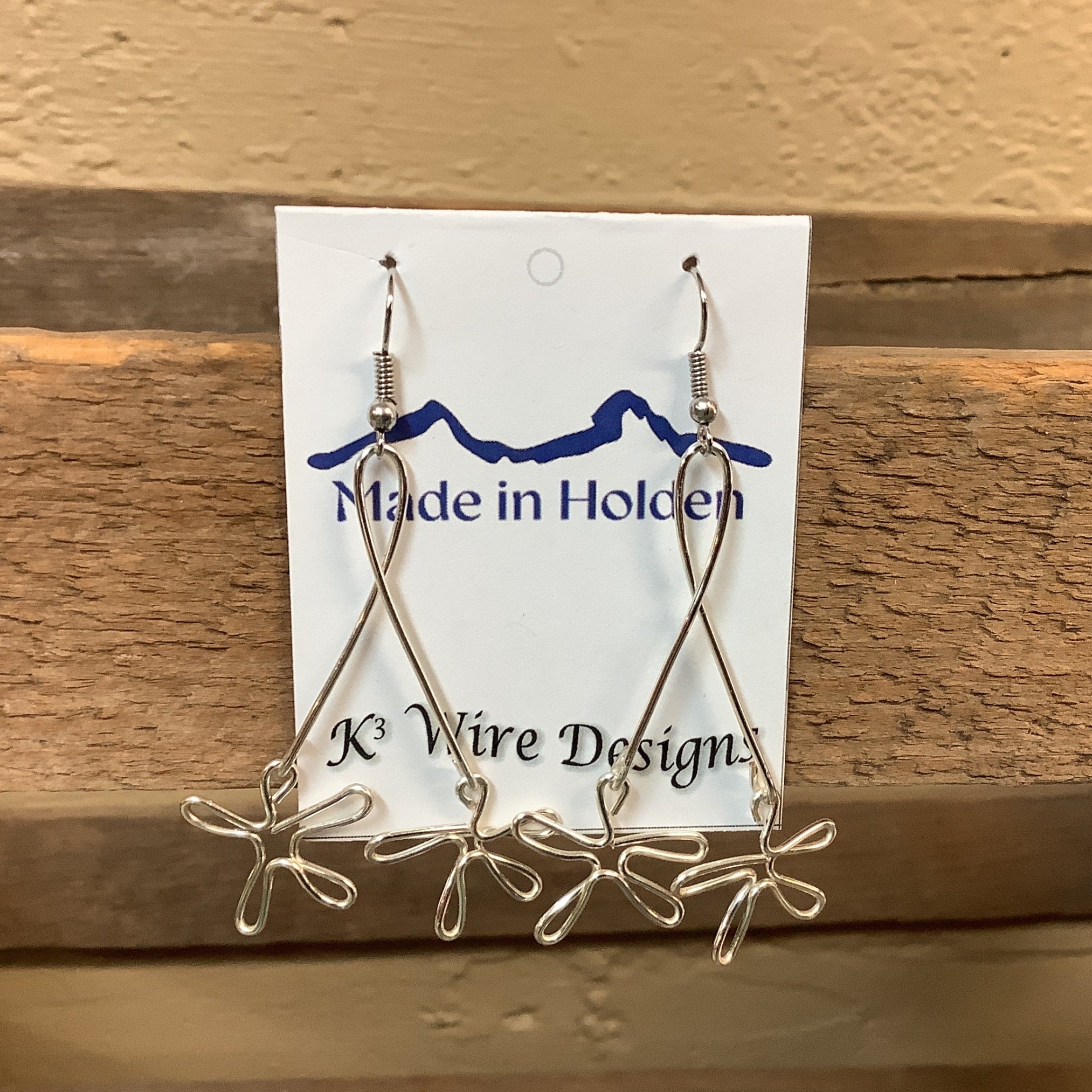 A pair of handmade silver wire dangle earrings displayed on a white backing card that reads 'Made in Holden' and 'K3 Wire Designs,' hanging on a rustic wooden shelf in an indoor setting. SKU 8087