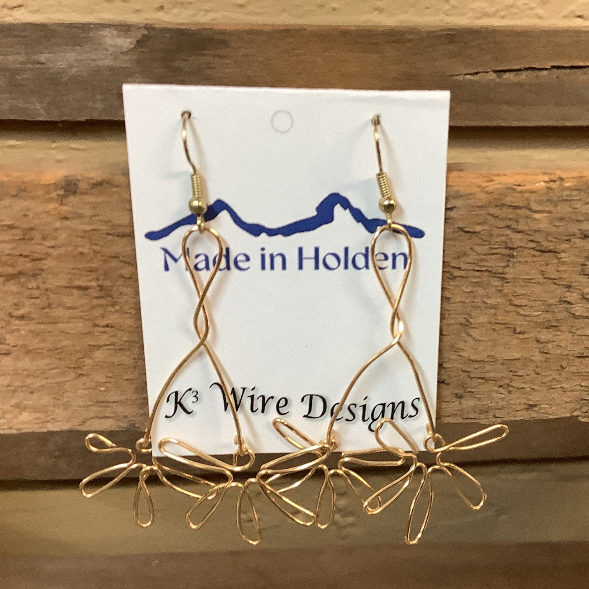A pair of handmade silver wire dangle earrings displayed on a white backing card that reads 'Made in Holden' and 'K3 Wire Designs,' hanging on a rustic wooden shelf in an indoor setting. SKU 8085