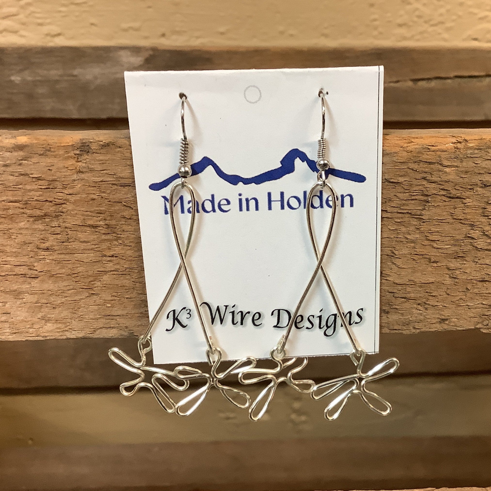 A pair of handmade silver wire dangle earrings displayed on a white backing card that reads 'Made in Holden' and 'K3 Wire Designs,' hanging on a rustic wooden shelf in an indoor setting. SKU 8086