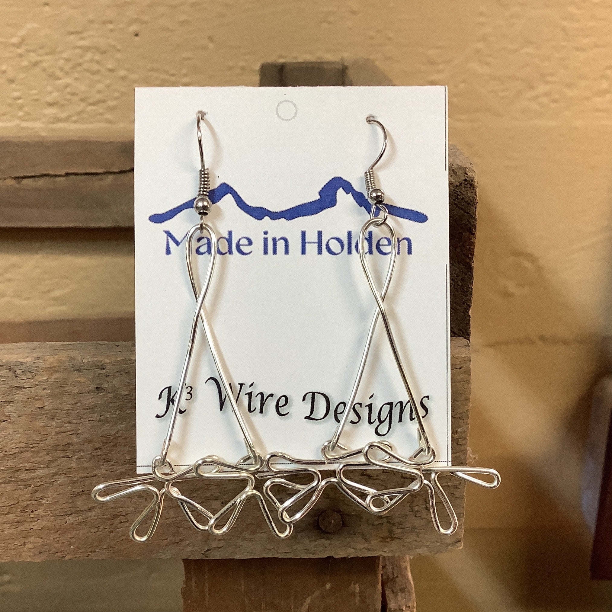 A pair of handmade silver wire dangle earrings displayed on a white backing card that reads 'Made in Holden' and 'K3 Wire Designs,' hanging on a rustic wooden shelf in an indoor setting. SKU 8090