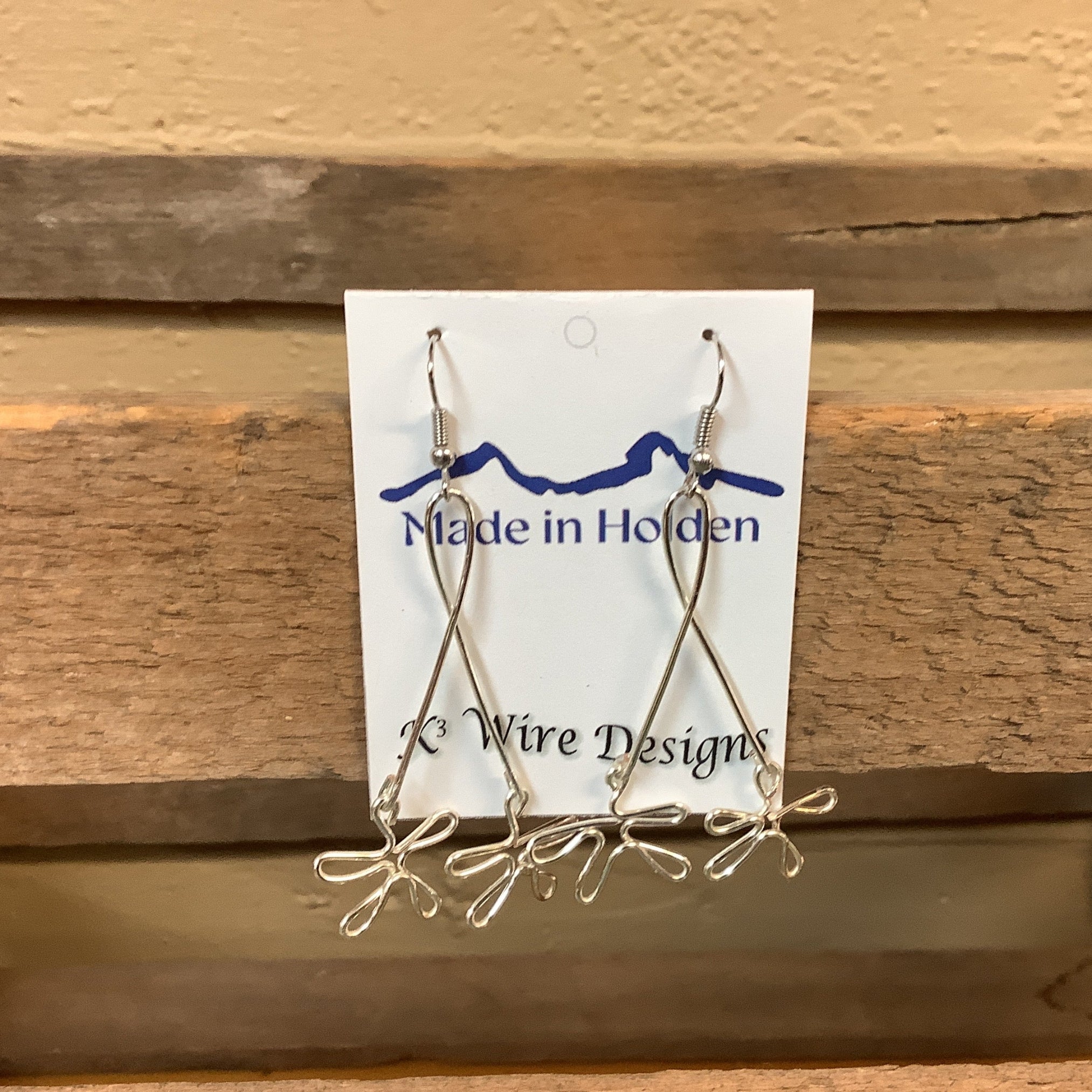A pair of handmade silver wire dangle earrings displayed on a white backing card that reads 'Made in Holden' and 'K3 Wire Designs,' hanging on a rustic wooden shelf in an indoor setting. SKU 8089