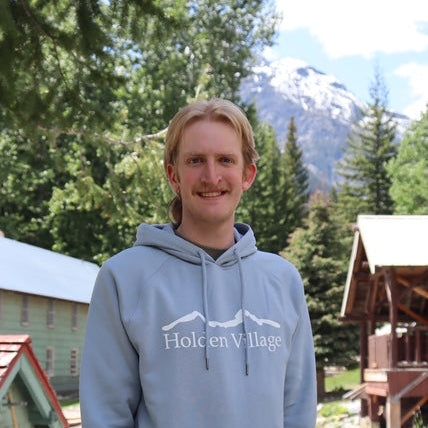 Person wearing a light blue hoodie with 'Holcen Village' logo outdoors with trees and mountains in the background