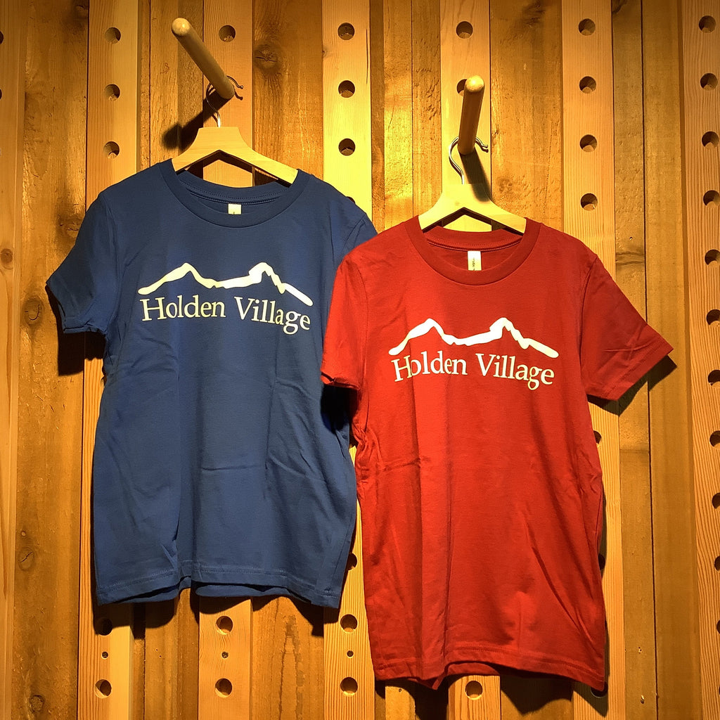 Two t-shirts, one red and one blue, hanging on a wooden pegboard. Both shirts have 'Hidden Village' printed in white script on the front with a white mountain graphic below.