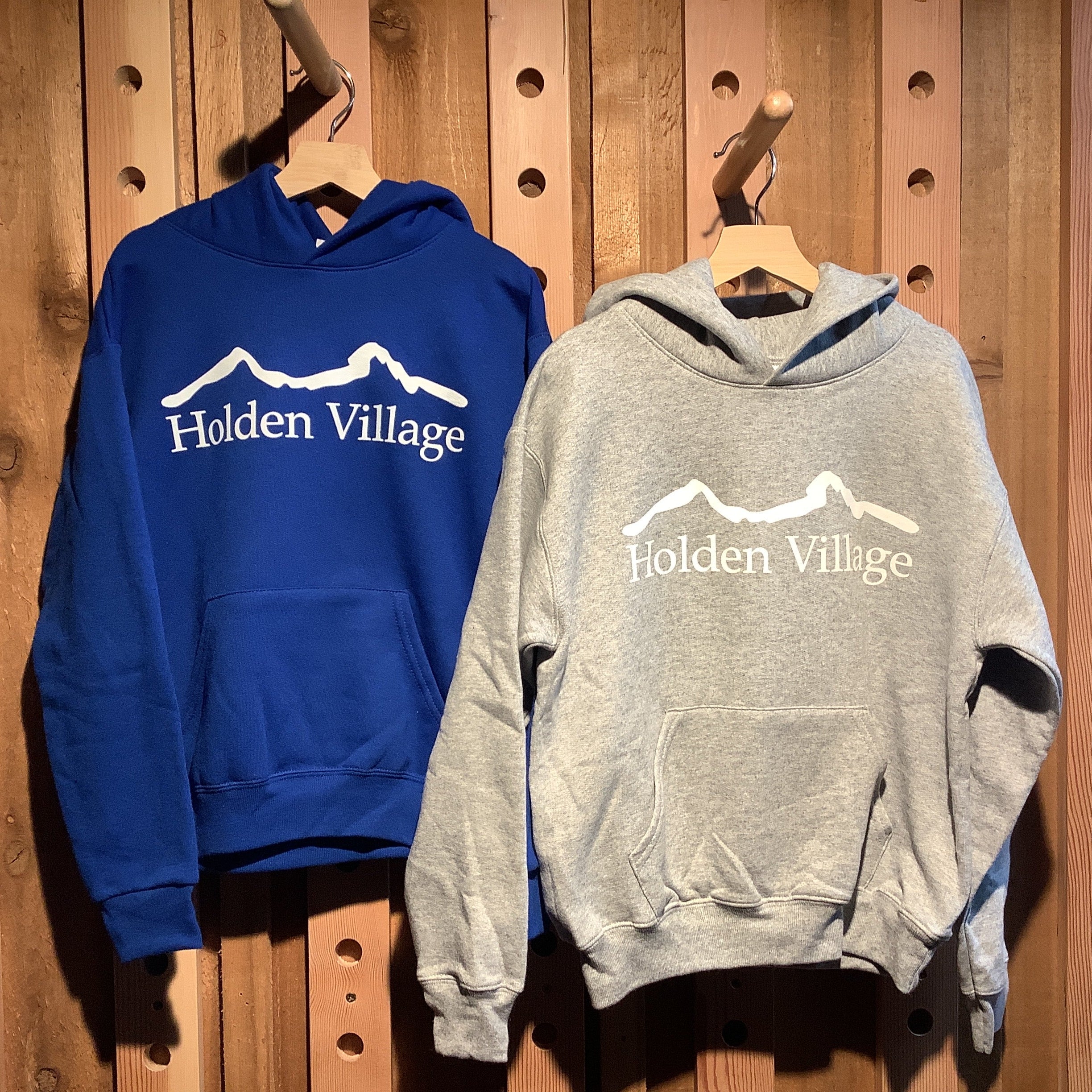 Two hoodies, one blue and one gray, hanging on a wooden pegboard with 'Holden Village' logo.