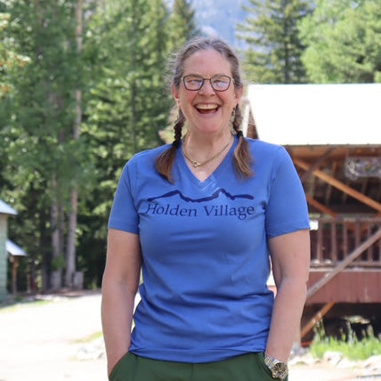 Person wearing a blue 'Holden Village' shirt standing outdoors with trees and mountains in the background