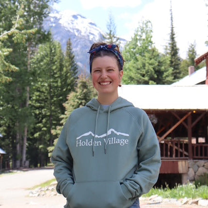 Person wearing a 'Holden Village' hoodie standing in front of a wooden building and trees.