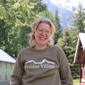 Person wearing a 'Holden Village' sweatshirt with a mountain design, standing outdoors with trees and buildings in the background.
