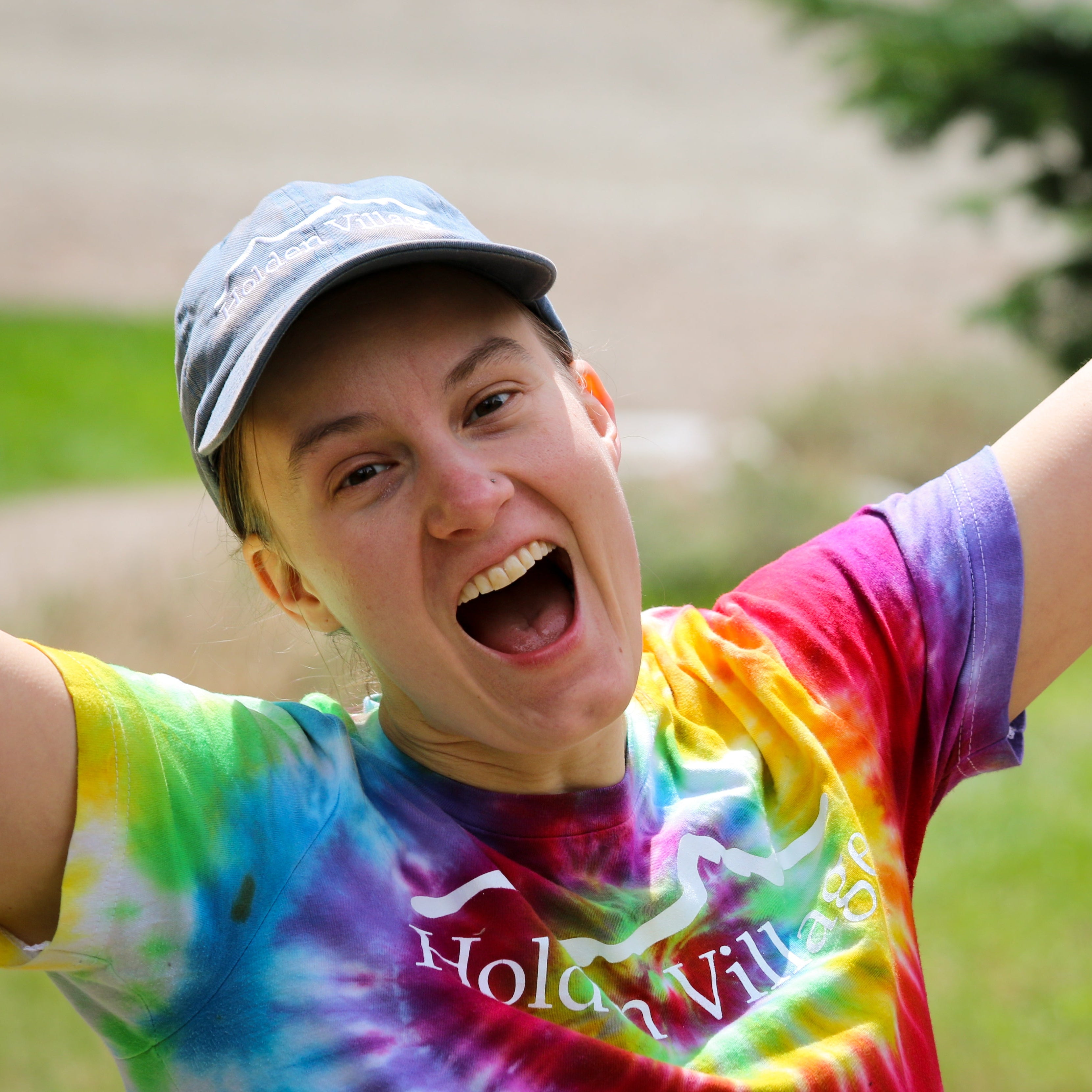 Person wearing a tie-dye shirt with 'Holden Village' text, outdoors with arms raised.