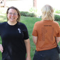 Two people standing outdoors, one wearing a black shirt with a logo and the other wearing an orange shirt with text.