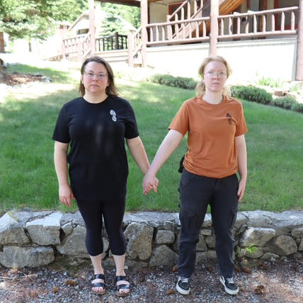 Two people standing outdoors in front of a house with a stone wall.