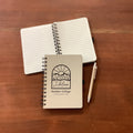 A small spiral-bound notebook with a white cover and black logo sits on top of an open lined notebook on a brown wooden table, with a silver pen beside it.