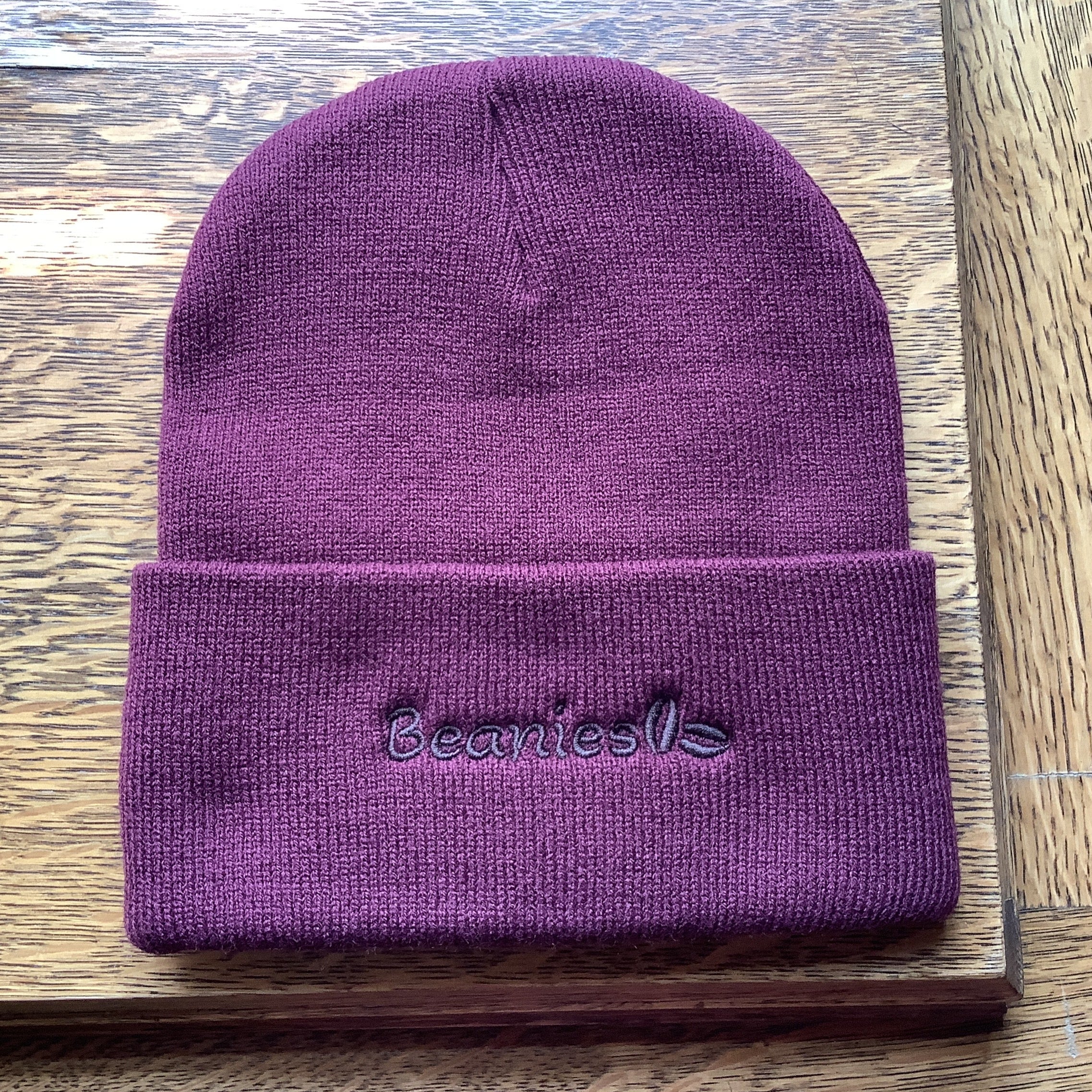 A maroon knit beanie hat with a folded cuff, featuring the word 'Beanies' embroidered in dark purple on the front, displayed on a wooden surface.