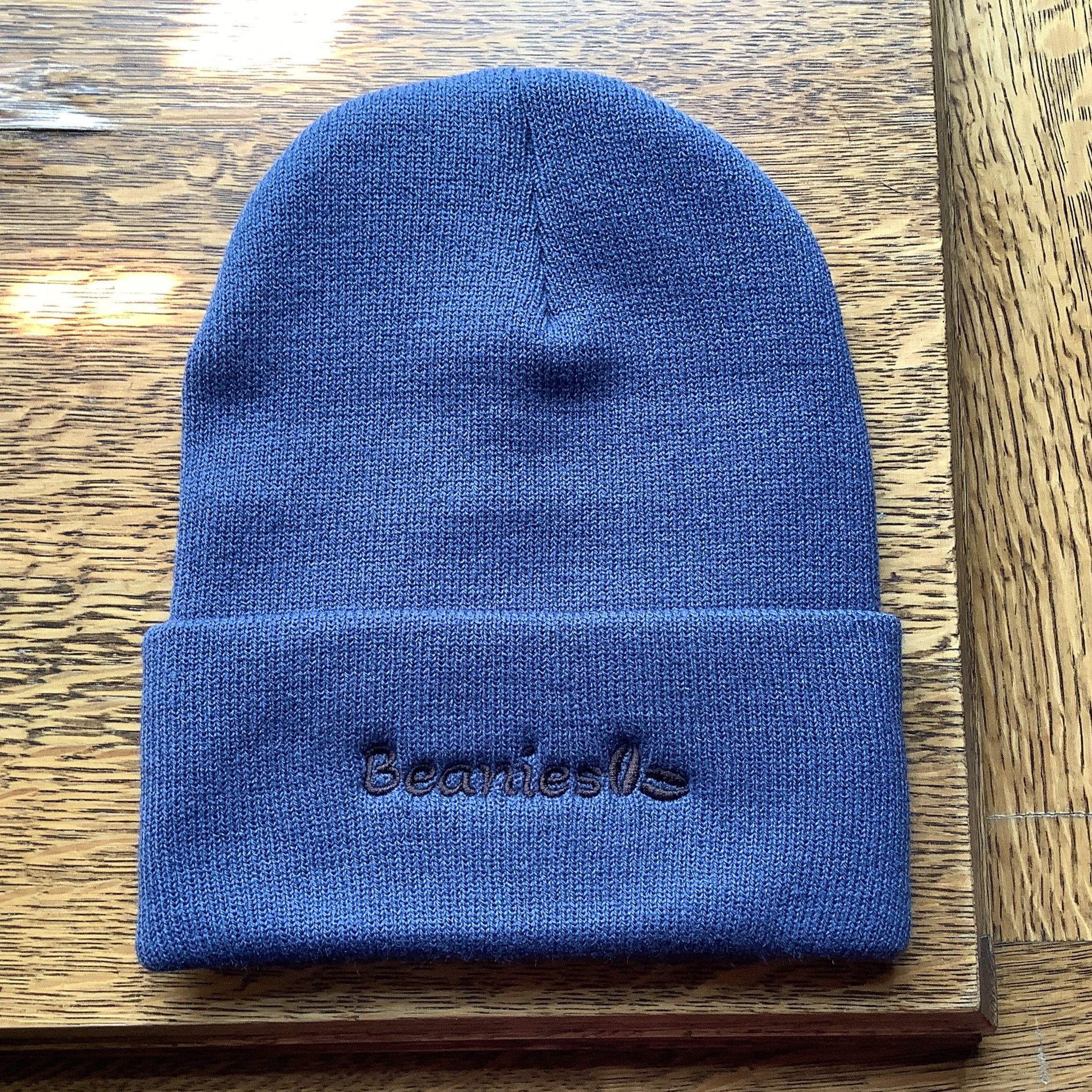 A blue knit beanie hat with a folded cuff, featuring the name 'Beanie' embroidered in black on the front, displayed on a wooden surface.