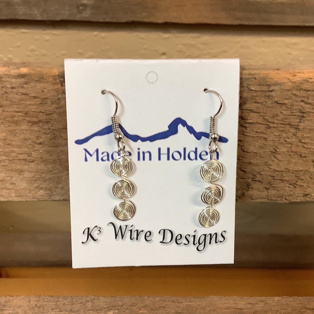 A pair of handmade silver wire spiral dangle earrings displayed on a white backing card that reads 'Made in Holden' and 'K3 Wire Designs,' hanging on a rustic wooden shelf in an indoor setting. SKU 7954