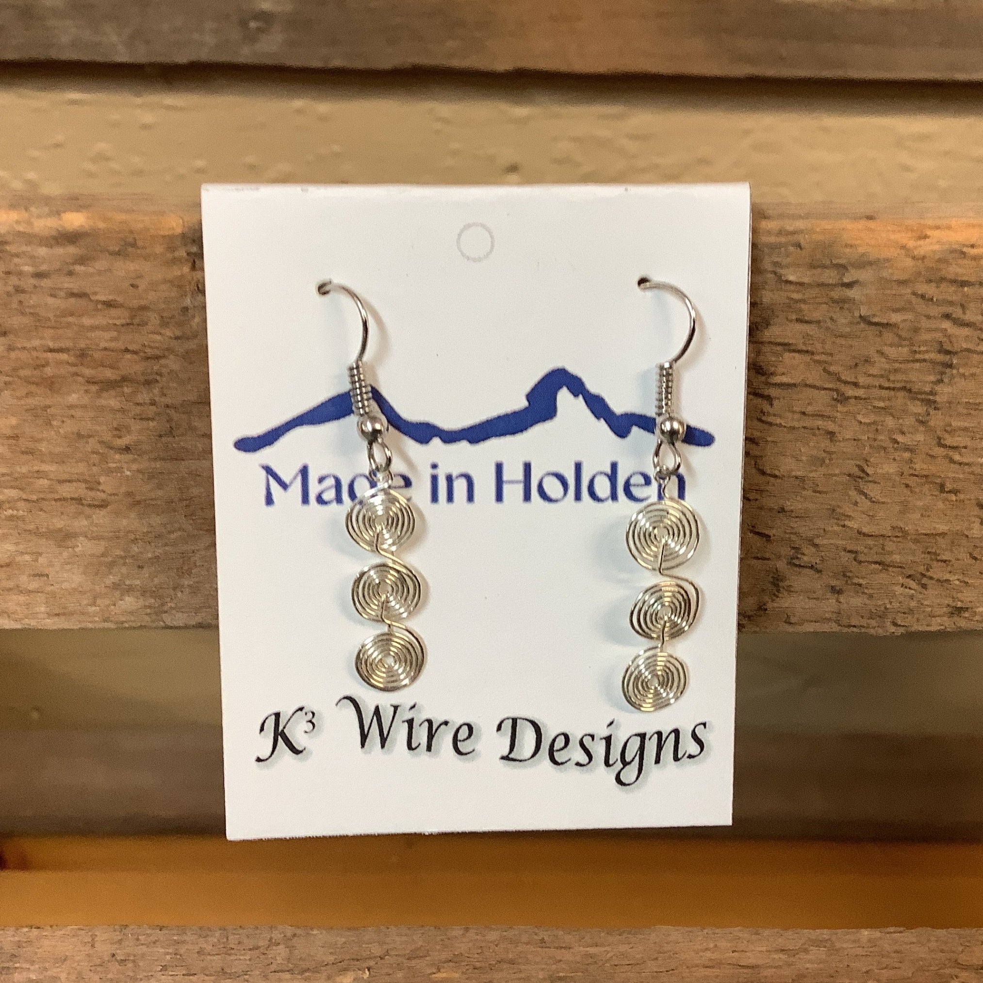 A pair of handmade silver wire spiral dangle earrings displayed on a white backing card that reads 'Made in Holden' and 'K3 Wire Designs,' hanging on a rustic wooden shelf in an indoor setting. SKU 7954