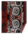 A rectangular woven blanket featuring a bold, stylized Indigenous-inspired graphic pattern in black, red, and light gray, with a fringed edge and a folded corner showing the textured weave.