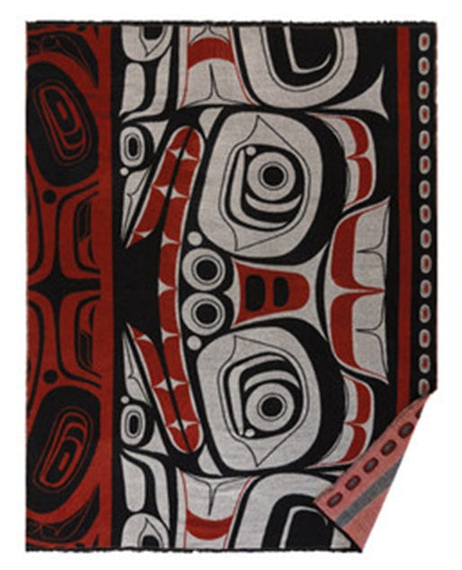A rectangular woven blanket featuring a bold, stylized Indigenous-inspired graphic pattern in black, red, and light gray, with a fringed edge and a folded corner showing the textured weave.