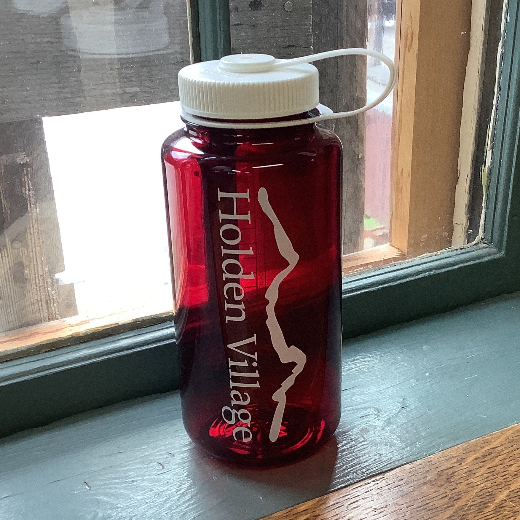 A red translucent Nalgene water bottle with a white cap and carrying loop, printed with 'Holden Village' and a white mountain graphic.