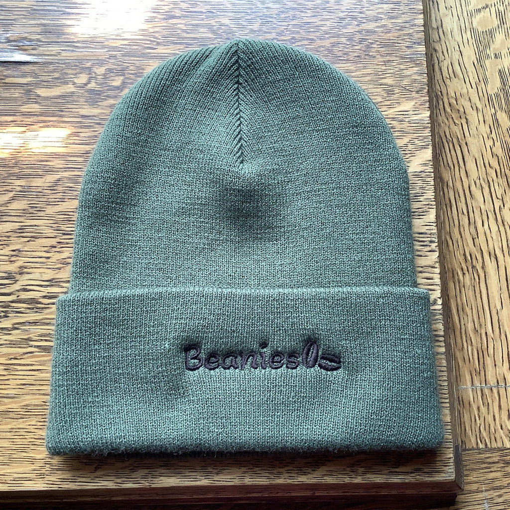 An olive green knit beanie with 'Beanies' embroidered in black on the front cuff, displayed on a wooden surface.