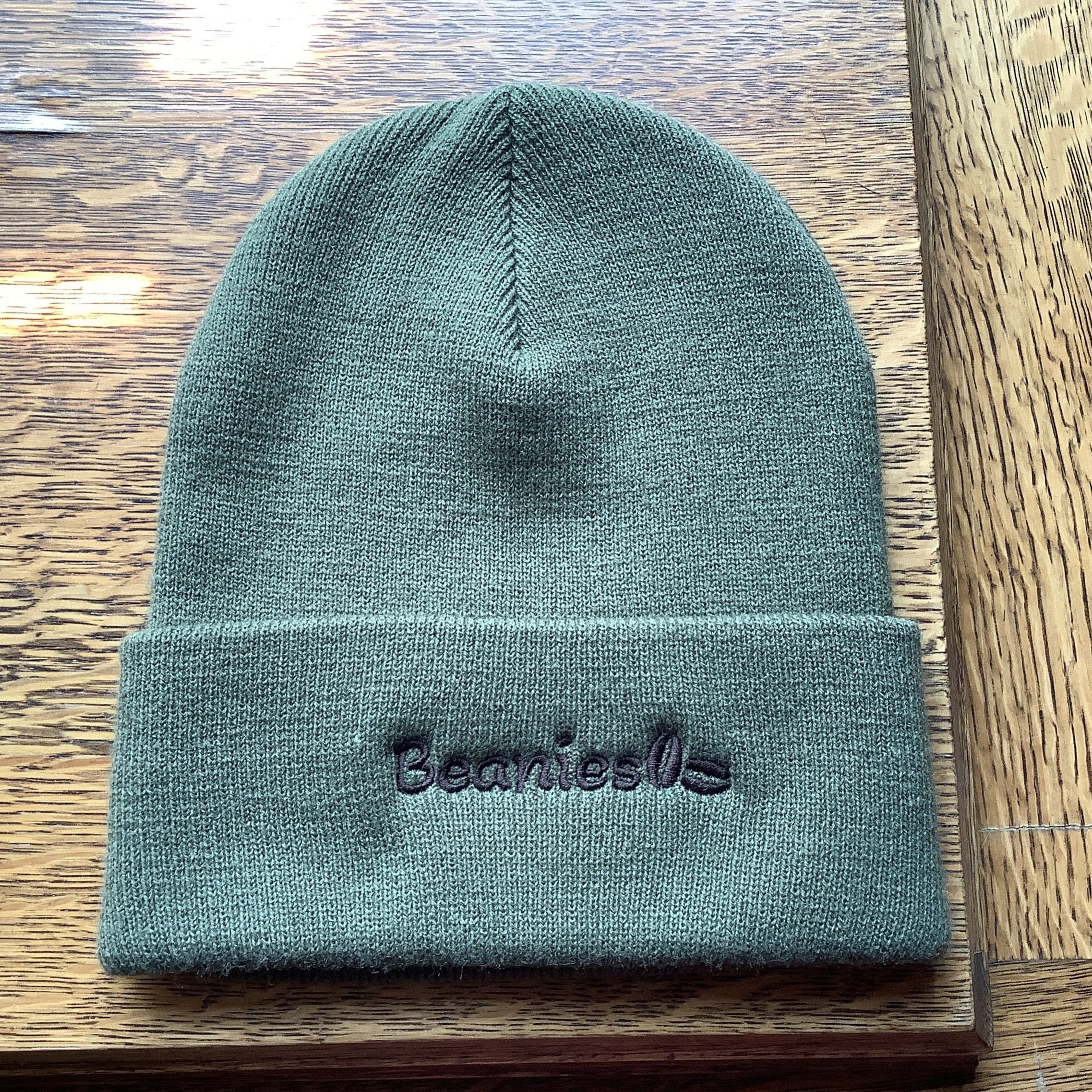 An olive green knit beanie with 'Beanies' embroidered in black on the front cuff, displayed on a wooden surface.