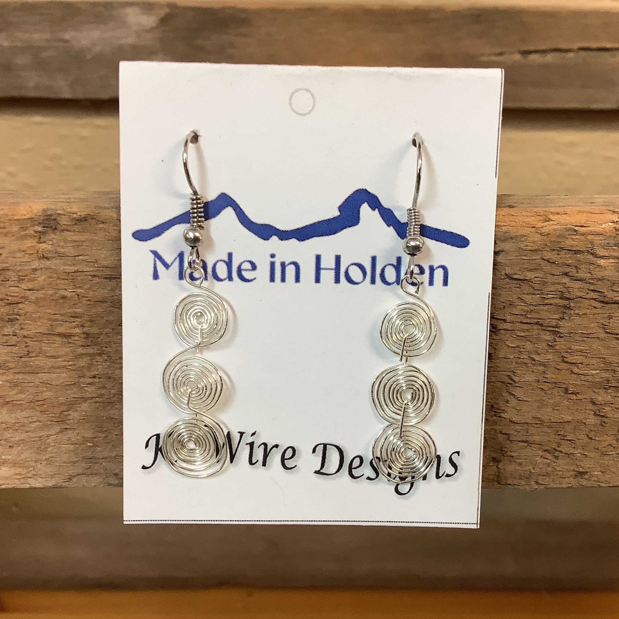 A pair of handmade silver wire spiral dangle earrings displayed on a white backing card that reads 'Made in Holden' and 'K3 Wire Designs,' hanging on a rustic wooden shelf in an indoor setting. SKU 8091